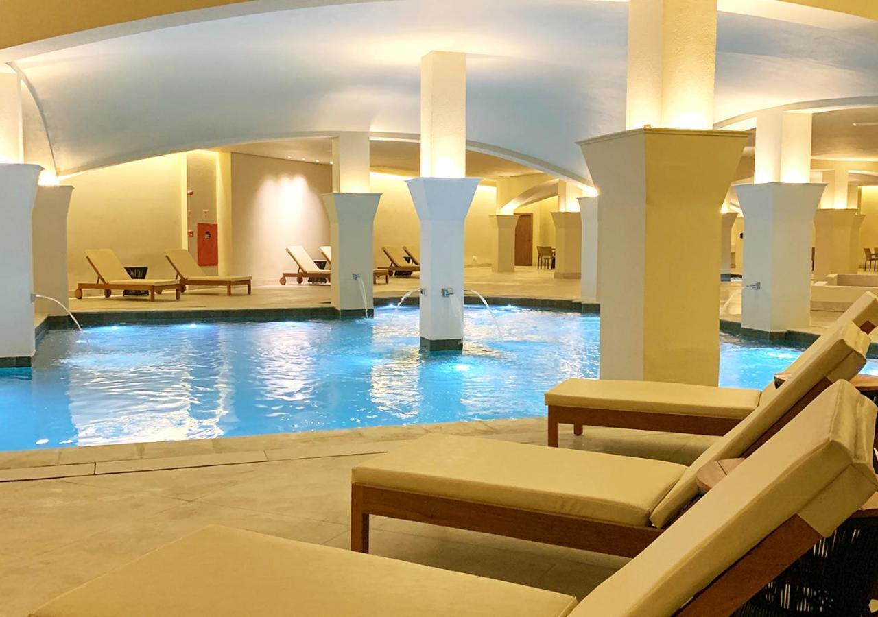 Buona Vitta Gramado Resort & Spa By Gramado Parks Екстериор снимка The photo shows an indoor spa or relaxation area with a tranquil atmosphere. There is a large swimming pool with clear blue water, illuminated by soft lighting. Surrounding the pool are several comfortable lounge chairs. The architecture features ele