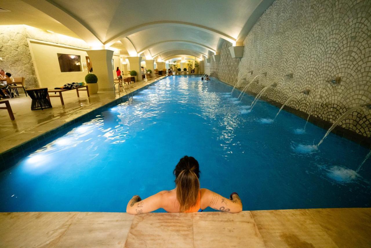 Buona Vitta Gramado Resort & Spa By Gramado Parks Екстериор снимка The photo shows an indoor swimming pool with a long, narrow design. The water is a clear blue, and there are features such as fountains along one side. In the foreground, a person with long hair is sitting on the edge of the pool, looking out over th
