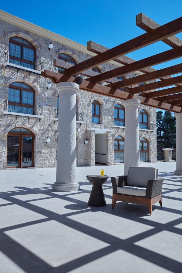 Buona Vitta Gramado Resort & Spa By Gramado Parks Екстериор снимка The photo shows a well-designed outdoor space with a stone wall building in the background. There are several tall columns that support a wooden pergola overhead, creating a shaded area. In this space, there is a stylish chair and a small table besid