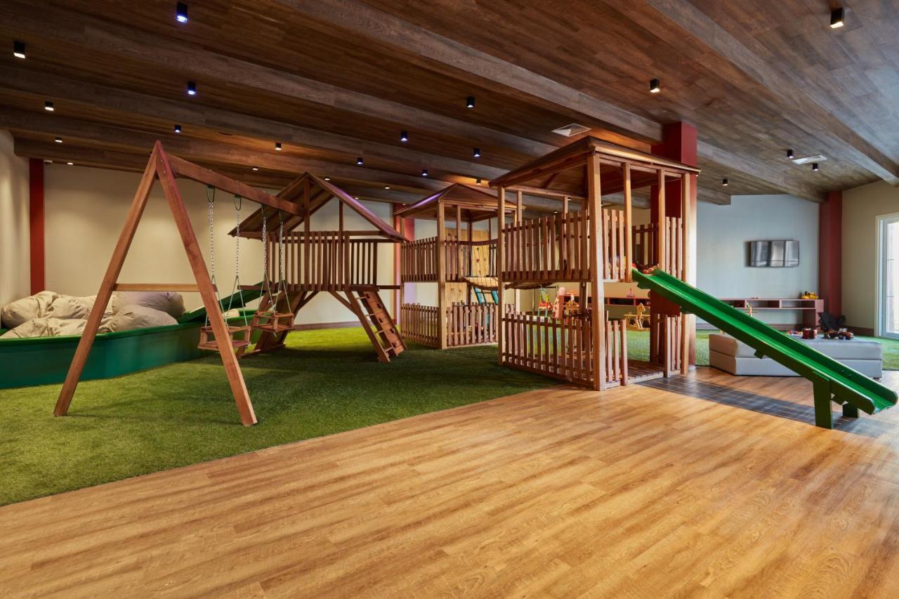 Buona Vitta Gramado Resort & Spa By Gramado Parks Екстериор снимка The photo shows an indoor play area designed for children. It features a wooden play structure that includes multiple levels, slides, and a swing set. The floor is covered with wooden planks, and there is a green carpet-like area that adds a soft tou