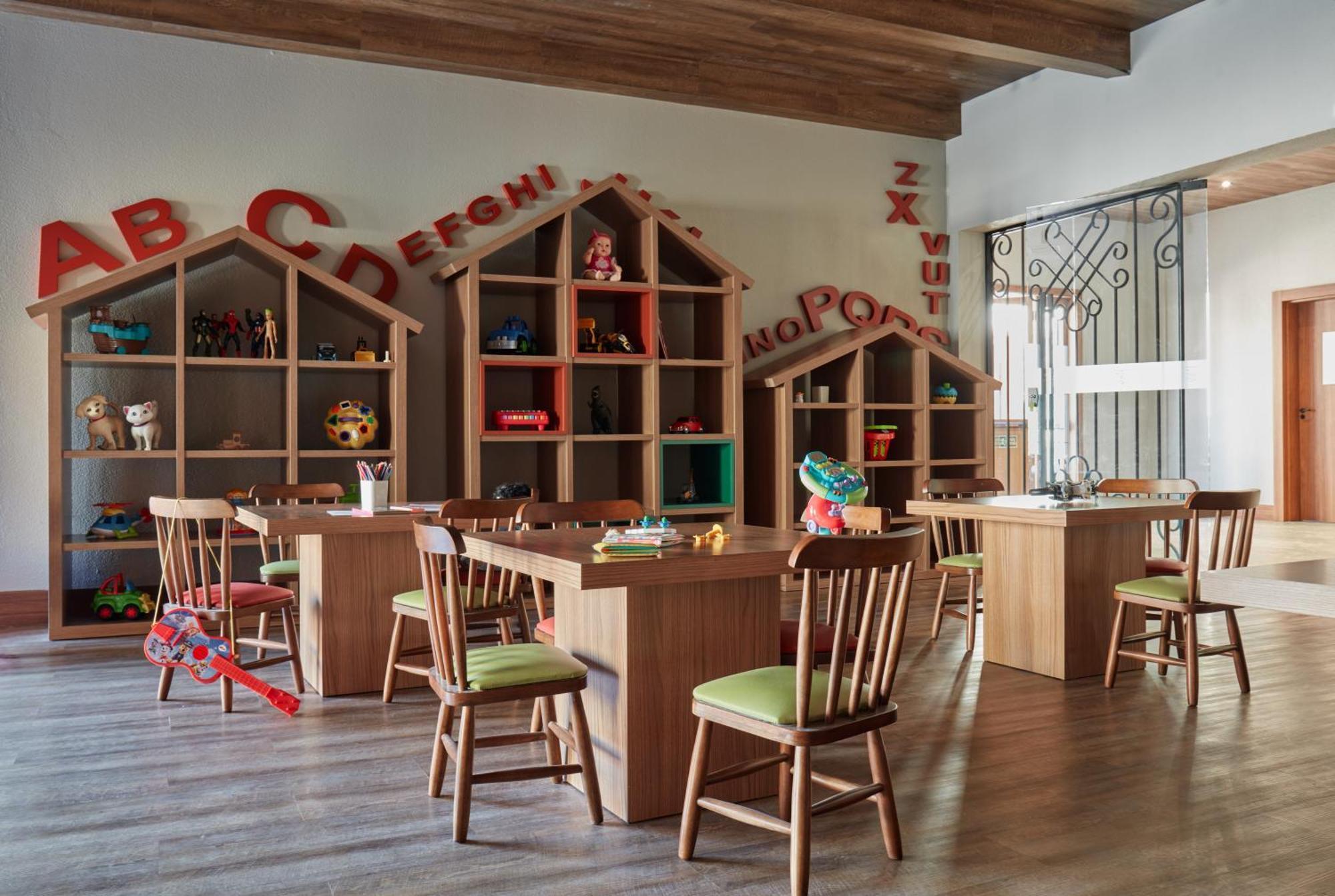 Buona Vitta Gramado Resort & Spa By Gramado Parks Екстериор снимка The photo shows a classroom or playroom designed for children. It features wooden furniture, including several tables and chairs with green cushions. In the background, there are large, open shelving units shaped like houses, displaying various toys 