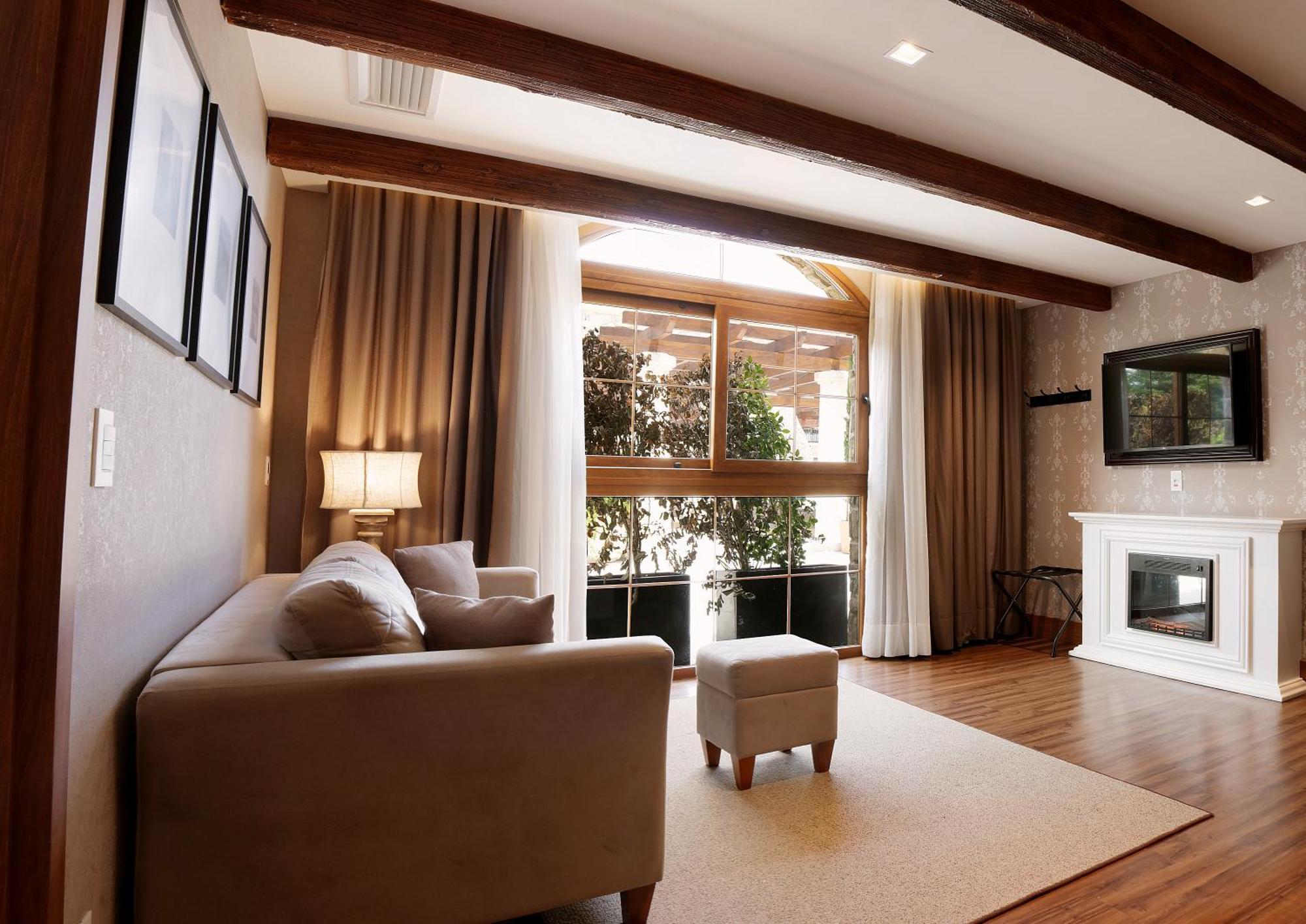 Buona Vitta Gramado Resort & Spa By Gramado Parks Екстериор снимка The photo depicts a cozy living room setting. It features a light-colored couch, a small ottoman, and a floor lamp beside the couch. Large windows are covered with curtains, allowing natural light to illuminate the space. In the background, there is 