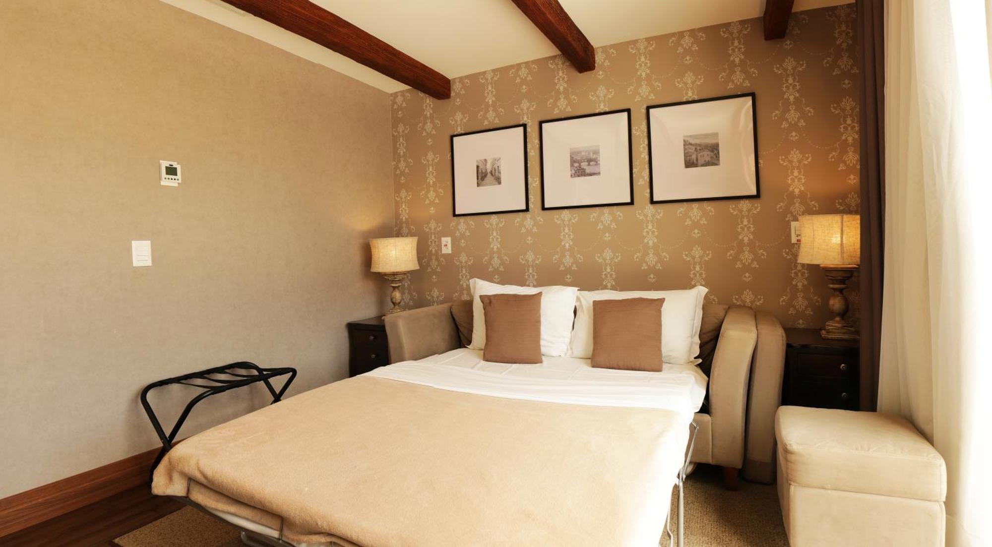 Buona Vitta Gramado Resort & Spa By Gramado Parks Екстериор снимка The photo shows a cozy bedroom with a neutral color palette. There is a bed with white linens and two brown decorative pillows. On either side of the bed, there are bedside tables with lamps. The walls feature a subtle floral pattern, and three frame