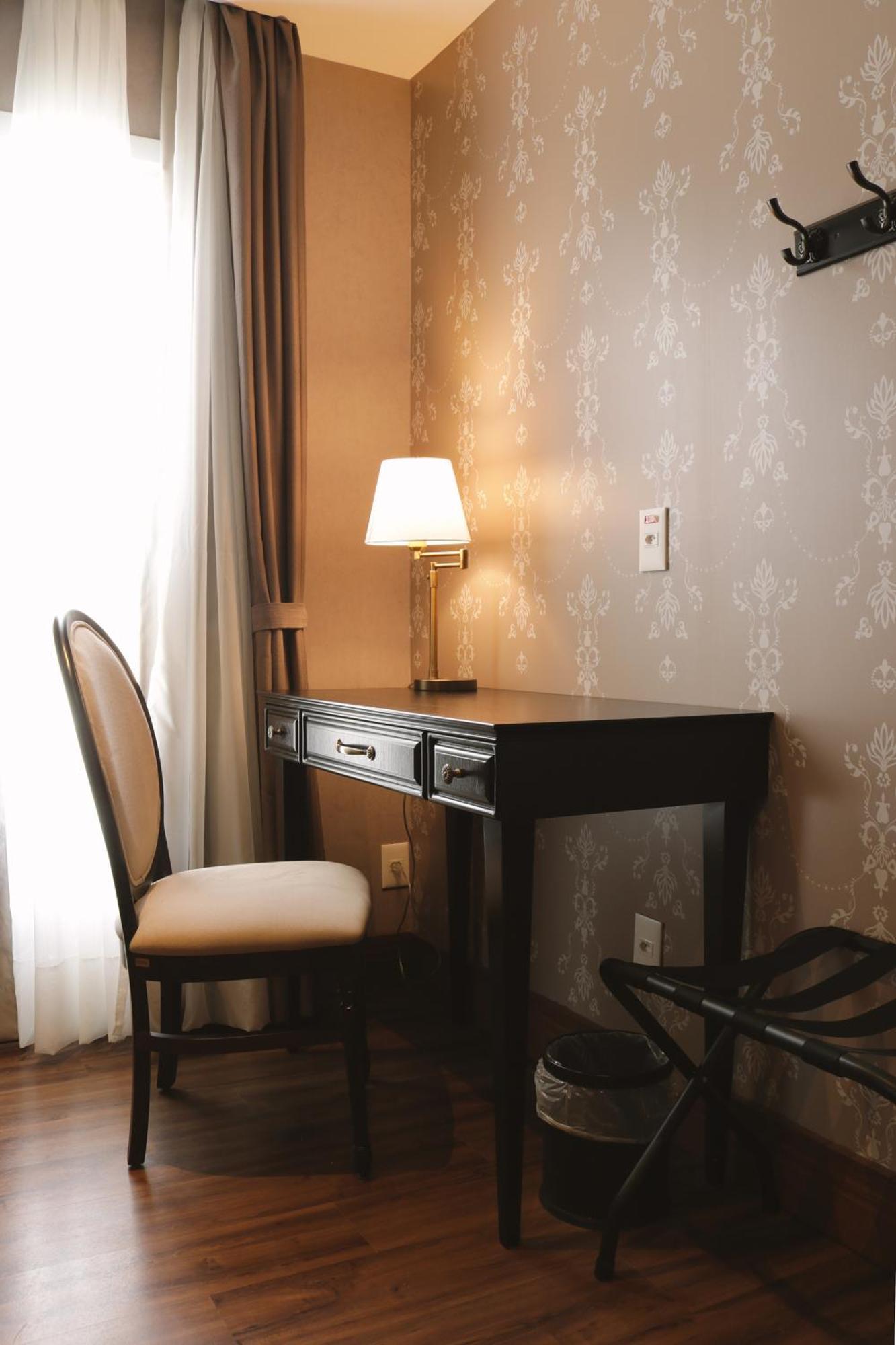 Buona Vitta Gramado Resort & Spa By Gramado Parks Екстериор снимка The photo shows a small desk set against a wall with a decorative pattern. The desk is black with a few drawers and has a small lamp on it. Next to the desk, there is a chair with a light-colored cushion. Soft light is coming through a window, castin