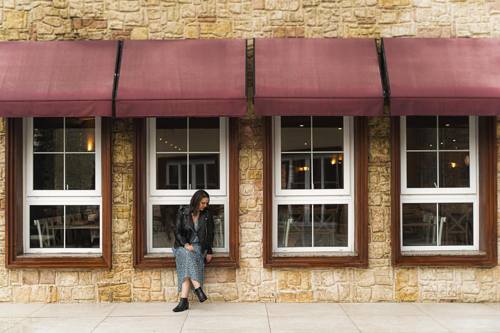Buona Vitta Gramado Resort & Spa By Gramado Parks Екстериор снимка The photo shows a woman standing against a stone wall with three large windows in front of her, each framed with wooden borders. The windows have maroon awnings above them. The woman is dressed in a long skirt and leather jacket, leaning slightly aga